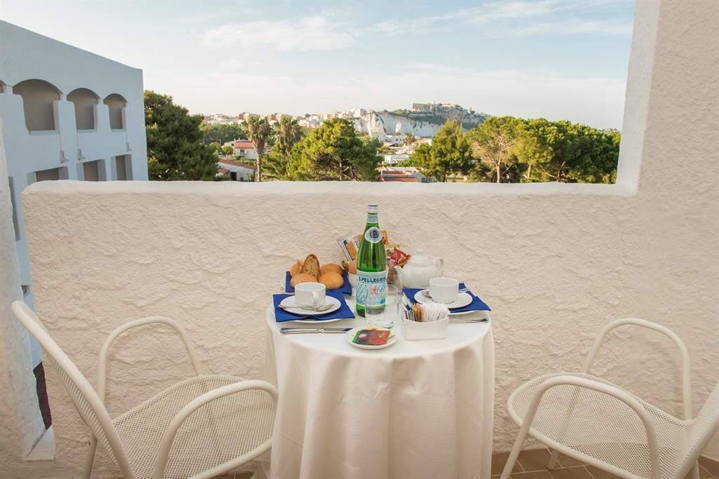 A balcony with a table set for breakfast, overlooking a scenic view of a coastal town.