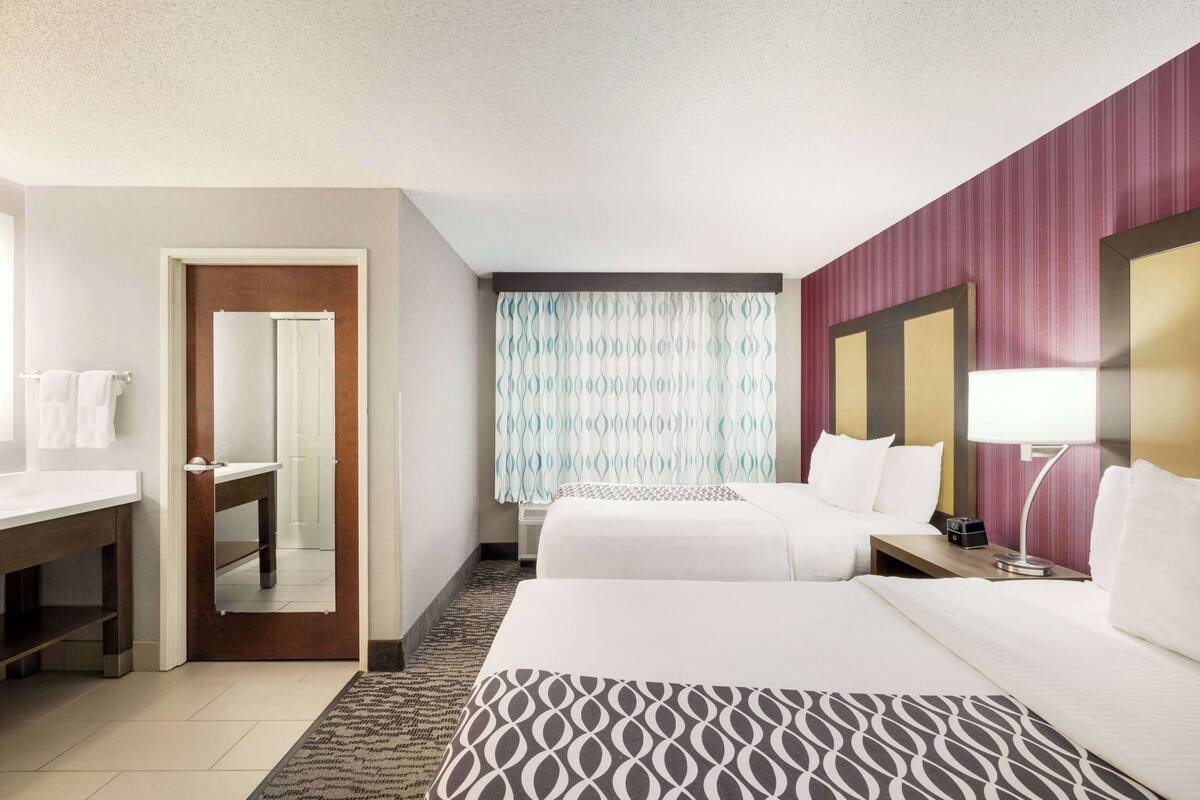 Une chambre d’hôtel avec deux lits, un bureau, un miroir, une fenêtre avec des rideaux à motifs et un mur orné de rayures.