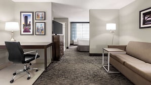 Habitación de hotel con escritorio, silla, sofá y televisor. Se observan cuadros en la pared y una ventana con cortinas.
