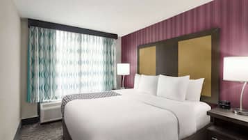 Una habitación de hotel con una cama grande, una mesita de noche con una lámpara, un televisor montado en la pared y un ventanal con cortinas estampadas.