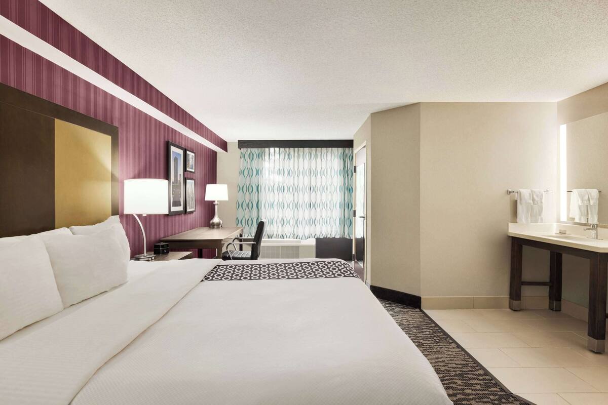 Une chambre d’hôtel avec un grand lit, un bureau avec une chaise, une lampe de bureau, une salle de bain avec un lavabo et un miroir, et un rideau à motifs.