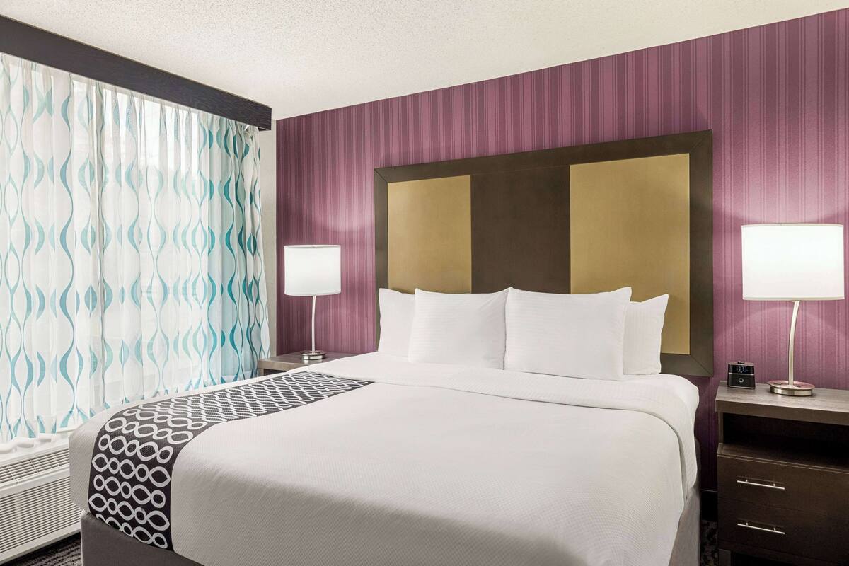 Une chambre d’hôtel avec un grand lit, deux tables de chevet avec des lampes, une tête de lit et une fenêtre avec des rideaux.