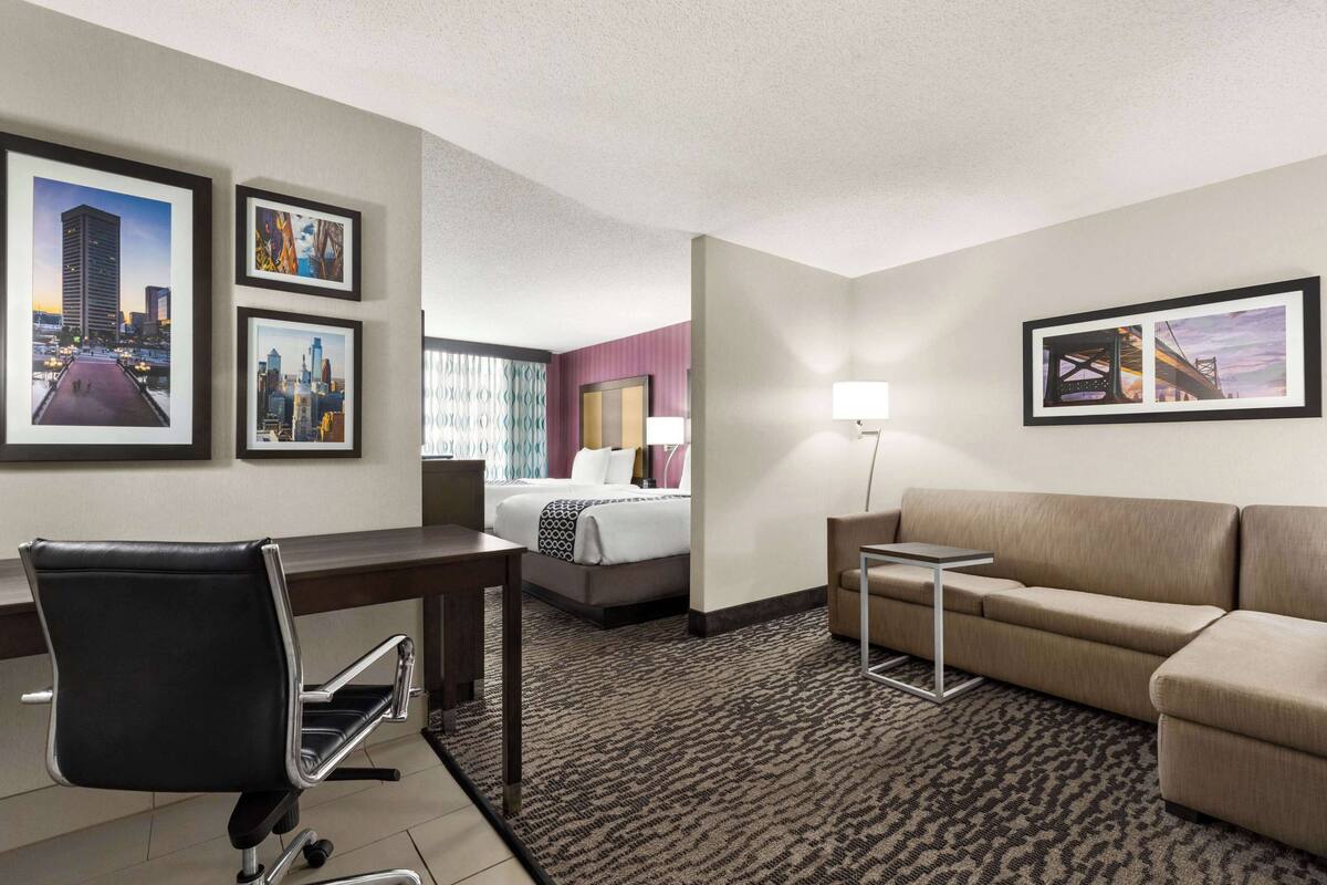 Une chambre d’hôtel avec un lit, un bureau, une chaise, un canapé et des tableaux encadrés aux murs.