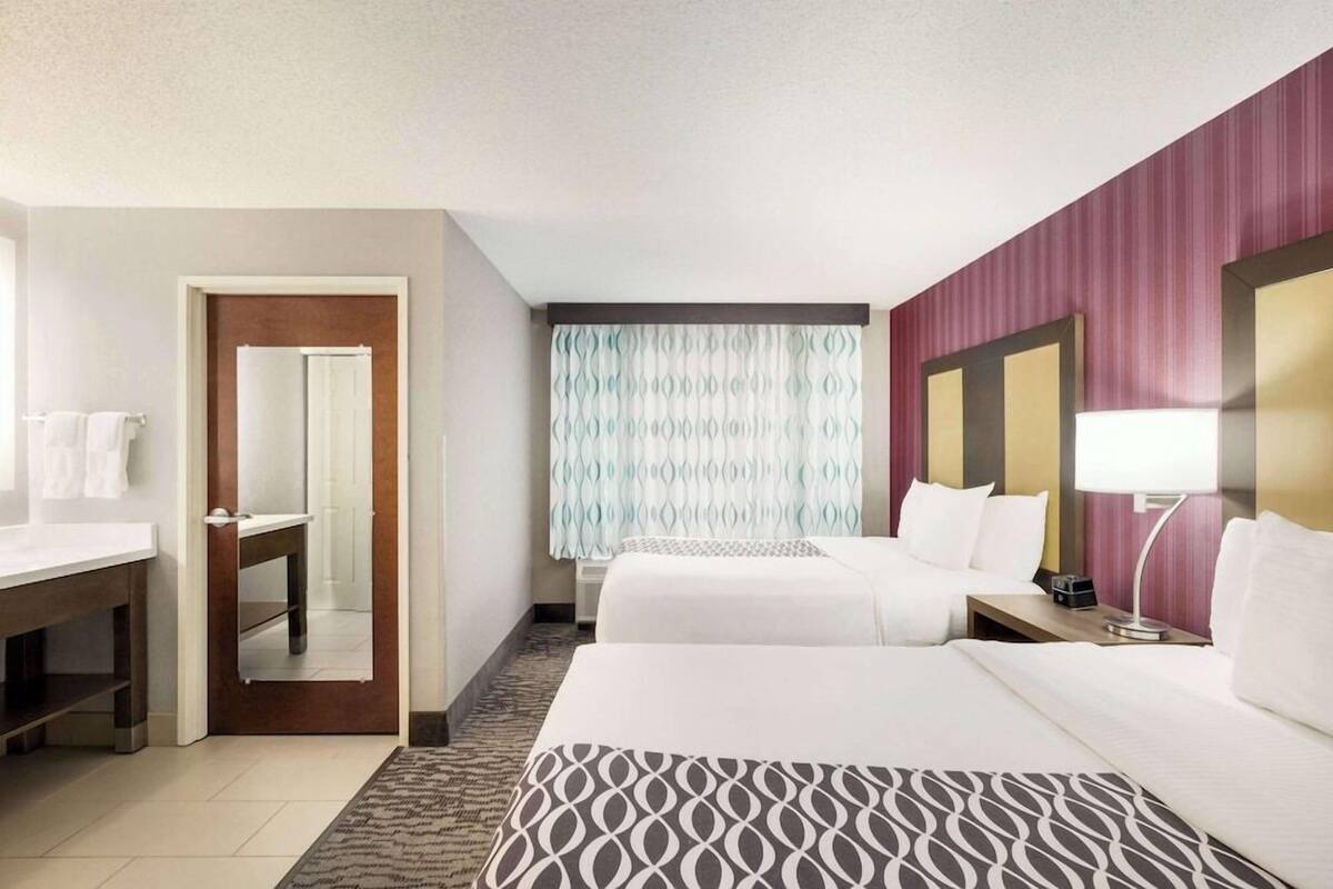 Une chambre d’hôtel avec deux lits, un bureau, un miroir, une fenêtre avec des rideaux à motifs et une lampe fixée au mur.
