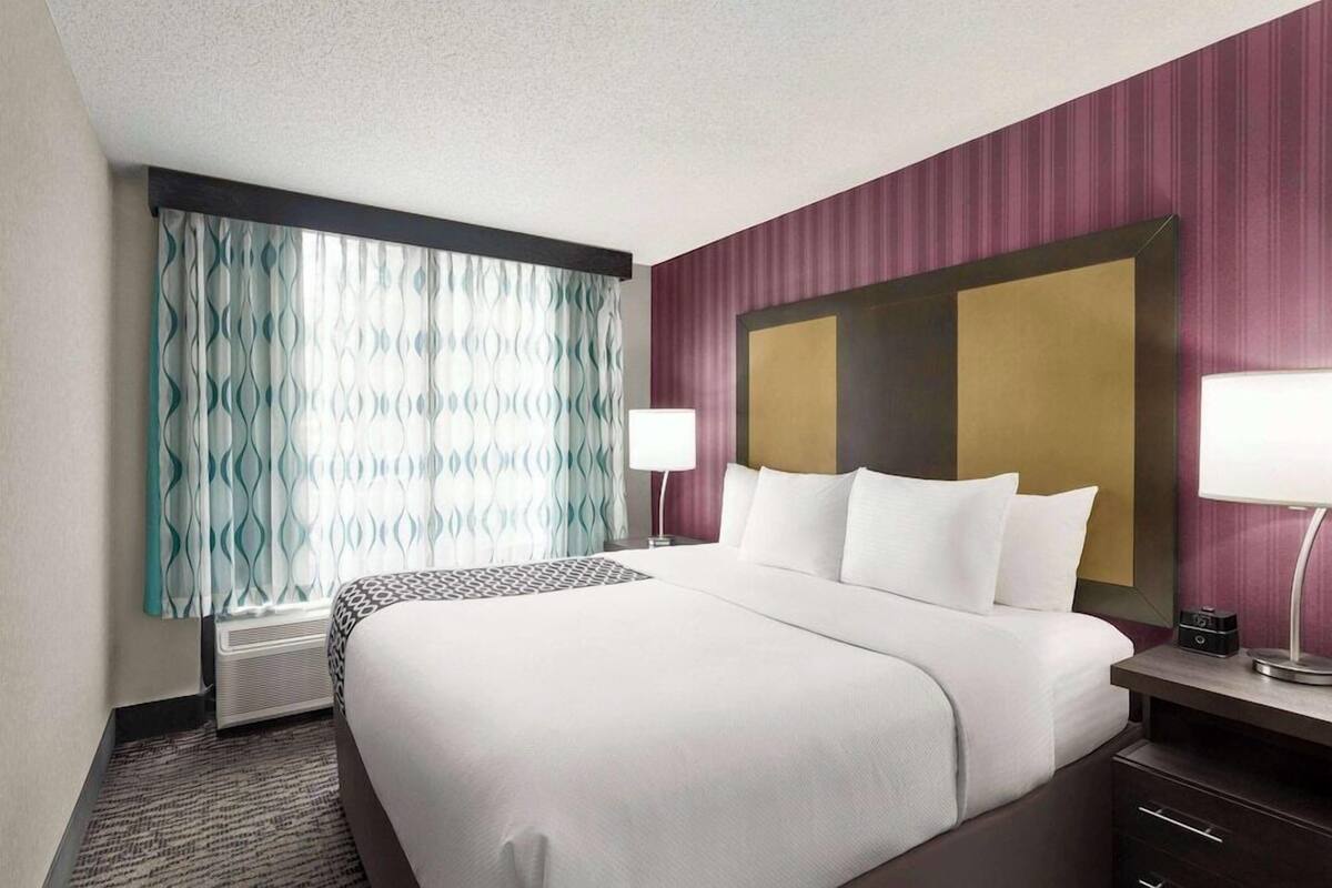 Une chambre d’hôtel avec un grand lit, une table de chevet, une lampe, une fenêtre avec des rideaux et un climatiseur fixé au mur.