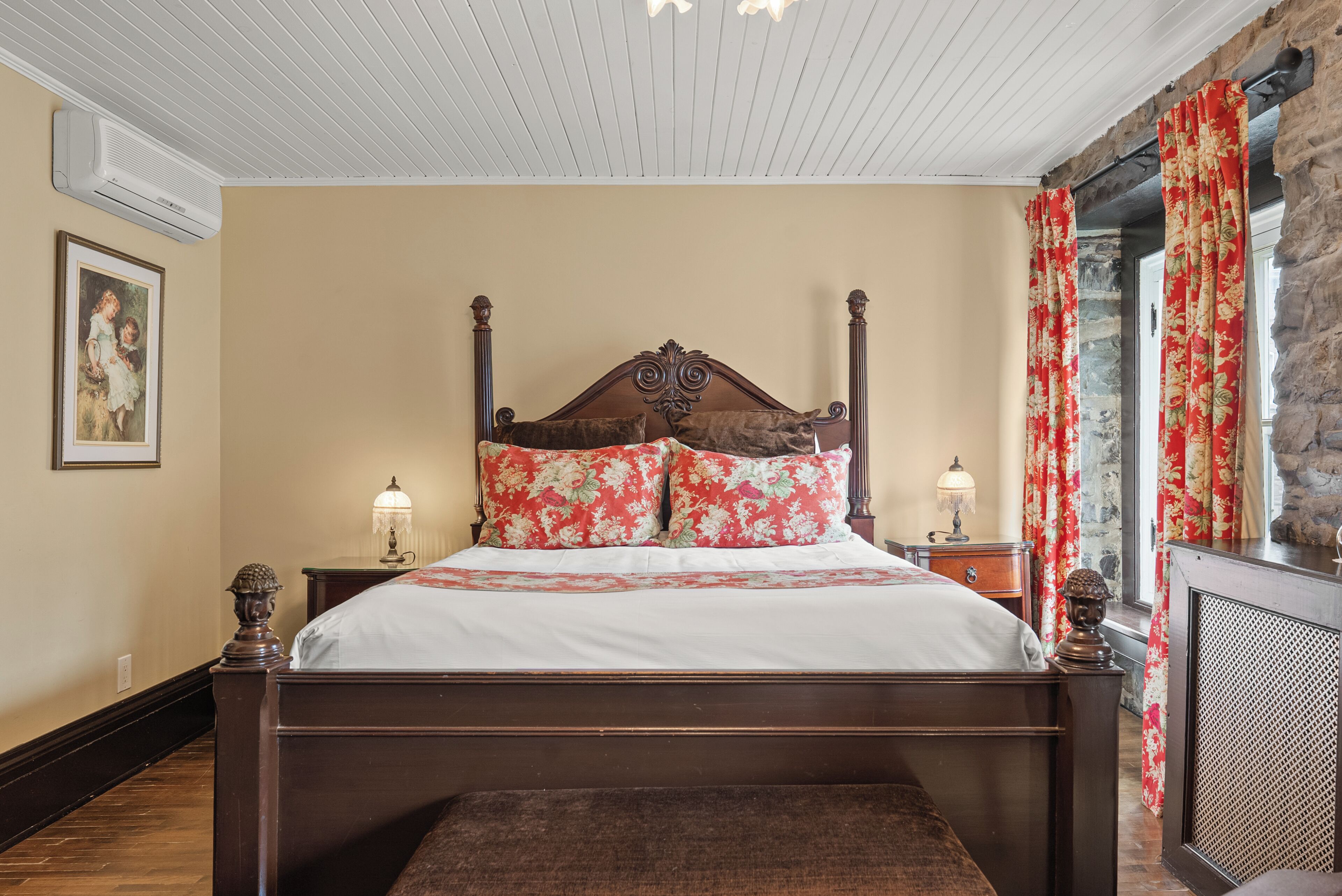 A bedroom with a wooden four-poster bed, floral pillows, a white bedspread, a nightstand with a lamp, and a stone wall.