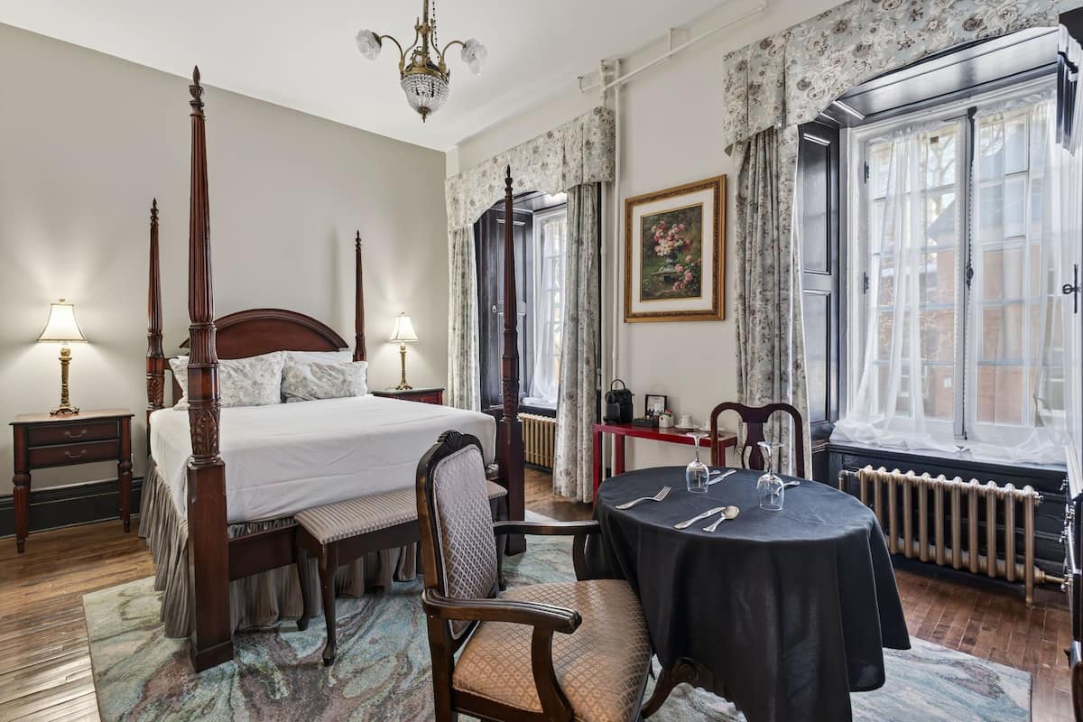 Una cama con dosel, una mesa de comedor con sillas, una araña y una ventana con cortinas.