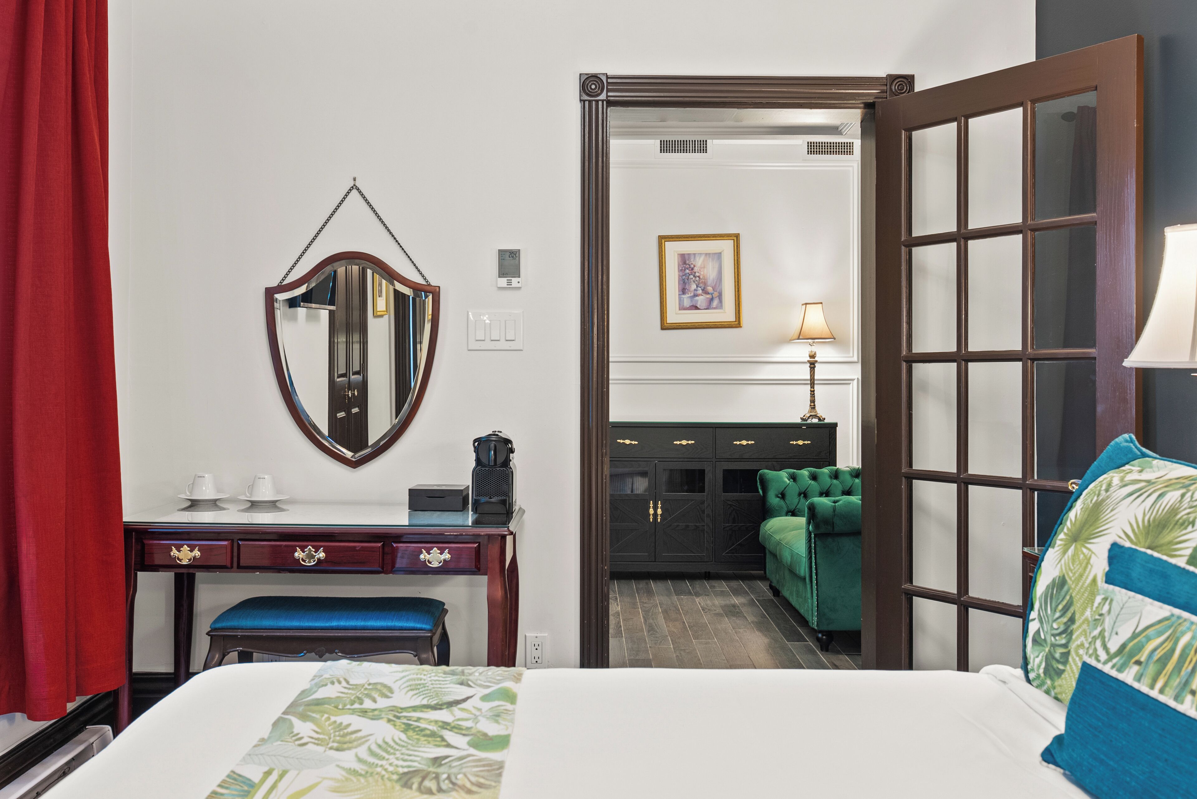 Une chambre d’hôtel avec un lit, un bureau doté d’un miroir, une commode, une chaise verte et une porte donnant sur un couloir.
