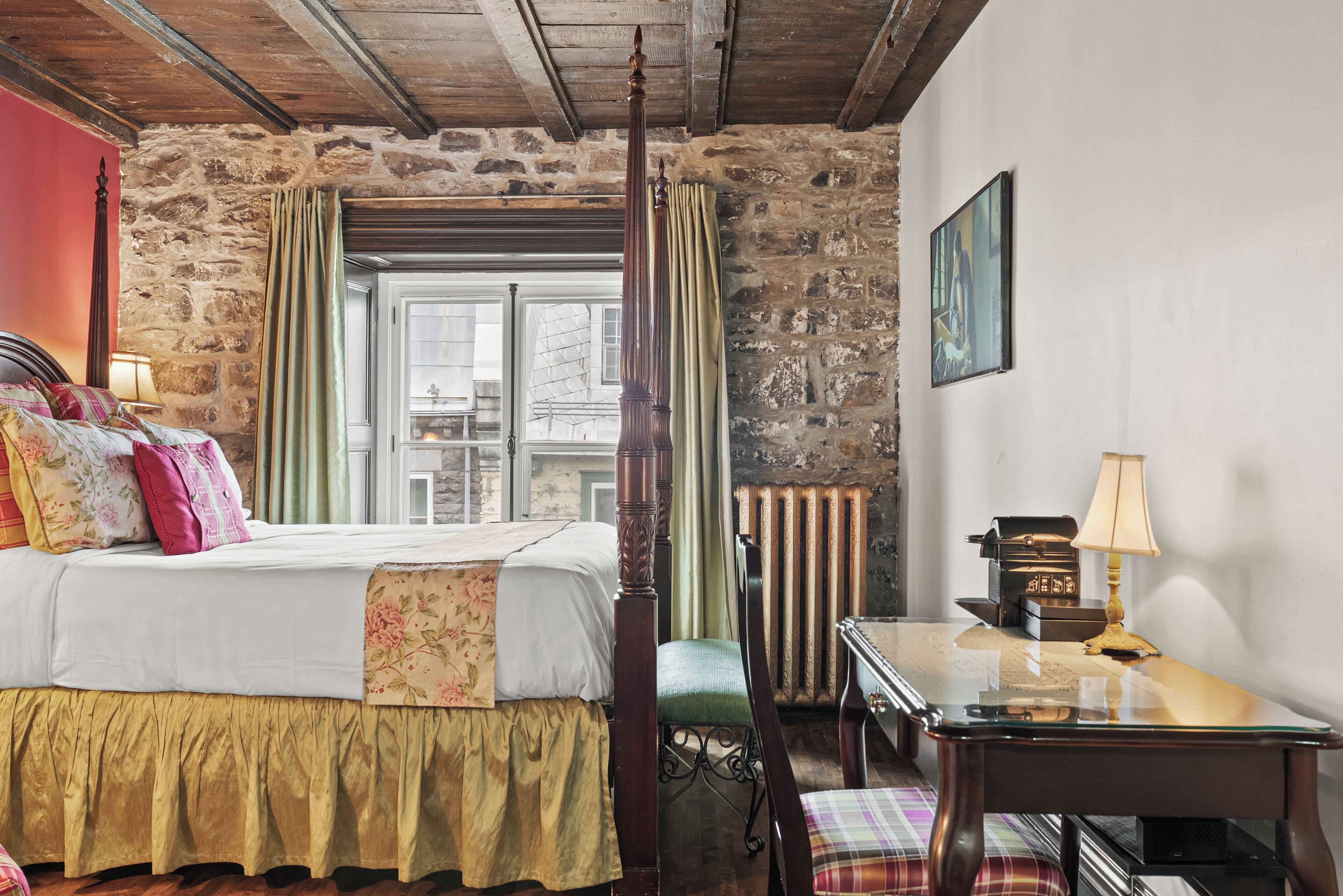 Ein Zimmer mit einem Himmelbett, einem Holztisch, einem Stuhl, einem Heizkörper und einem mit Vorhängen drapierten Fenster.