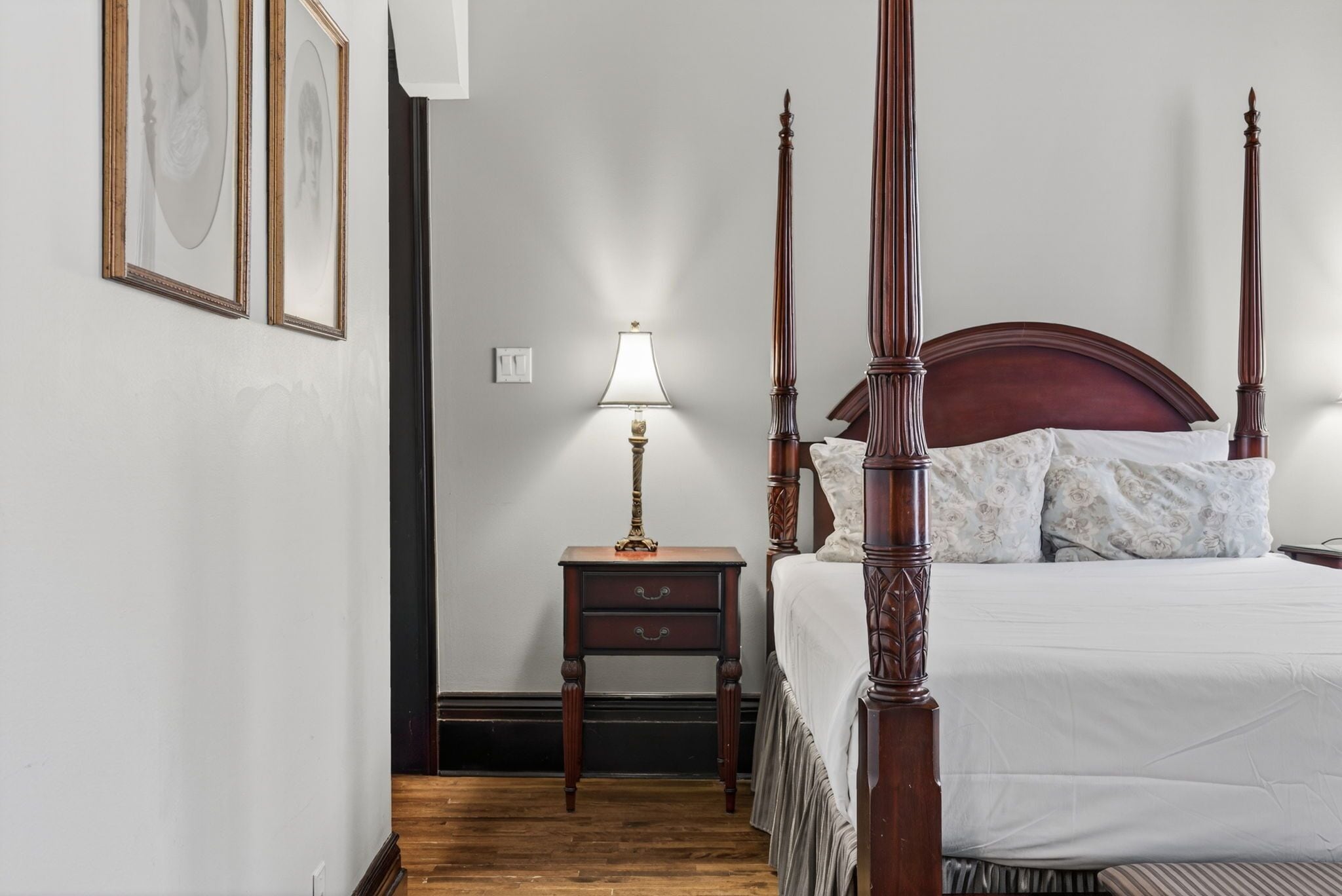 Una cama con dosel y tablero de madera, una mesita de noche con lámpara y cuadros enmarcados en la pared.