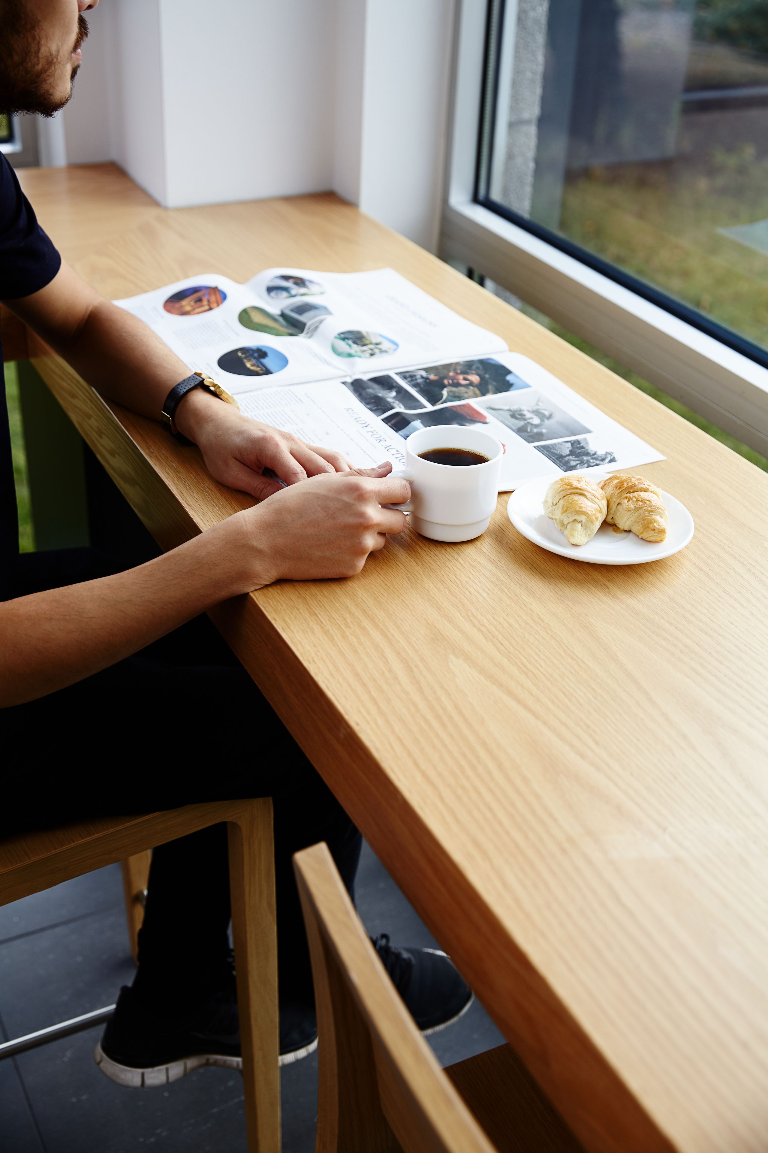 A person sitting at a wooden table with a cup of coffee and croissants, with a window in the background.