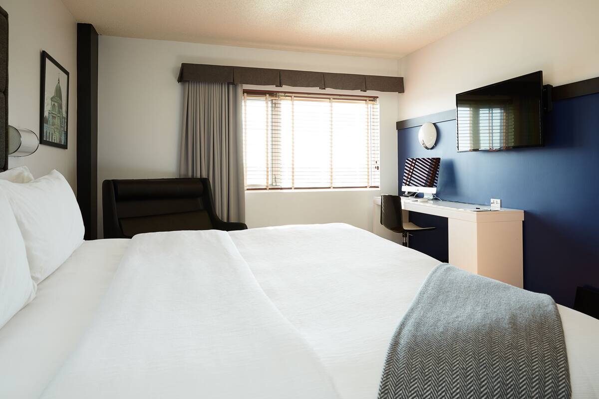 A hotel room with a large bed, a desk with a computer, a chair, and a window with blinds.