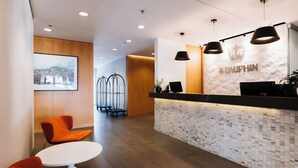 A reception area with a modern design, featuring a reception desk, seating area with orange chairs, and a large framed picture on the wall. - Hôtel Le Dauphin Montréal - Longueuil (Longueuil)