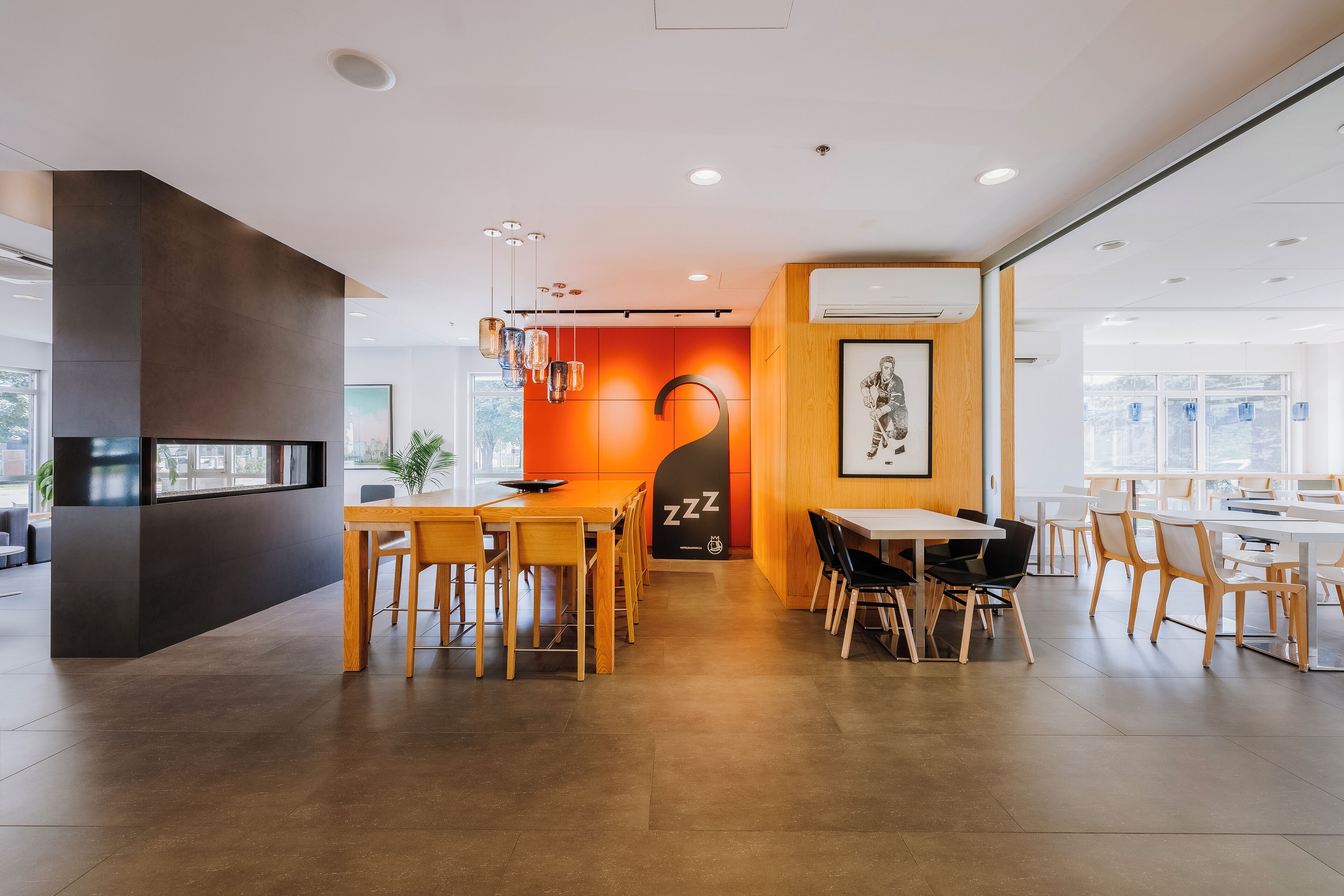 Un comedor moderno con mesas y sillas de madera, un amplio muro naranja y una obra de arte enmarcada.