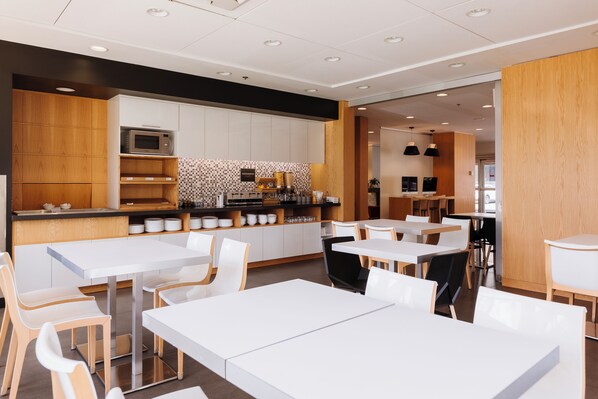 Un restaurante moderno con mesas y sillas blancas, una zona de cocina con microondas y detalles de madera.