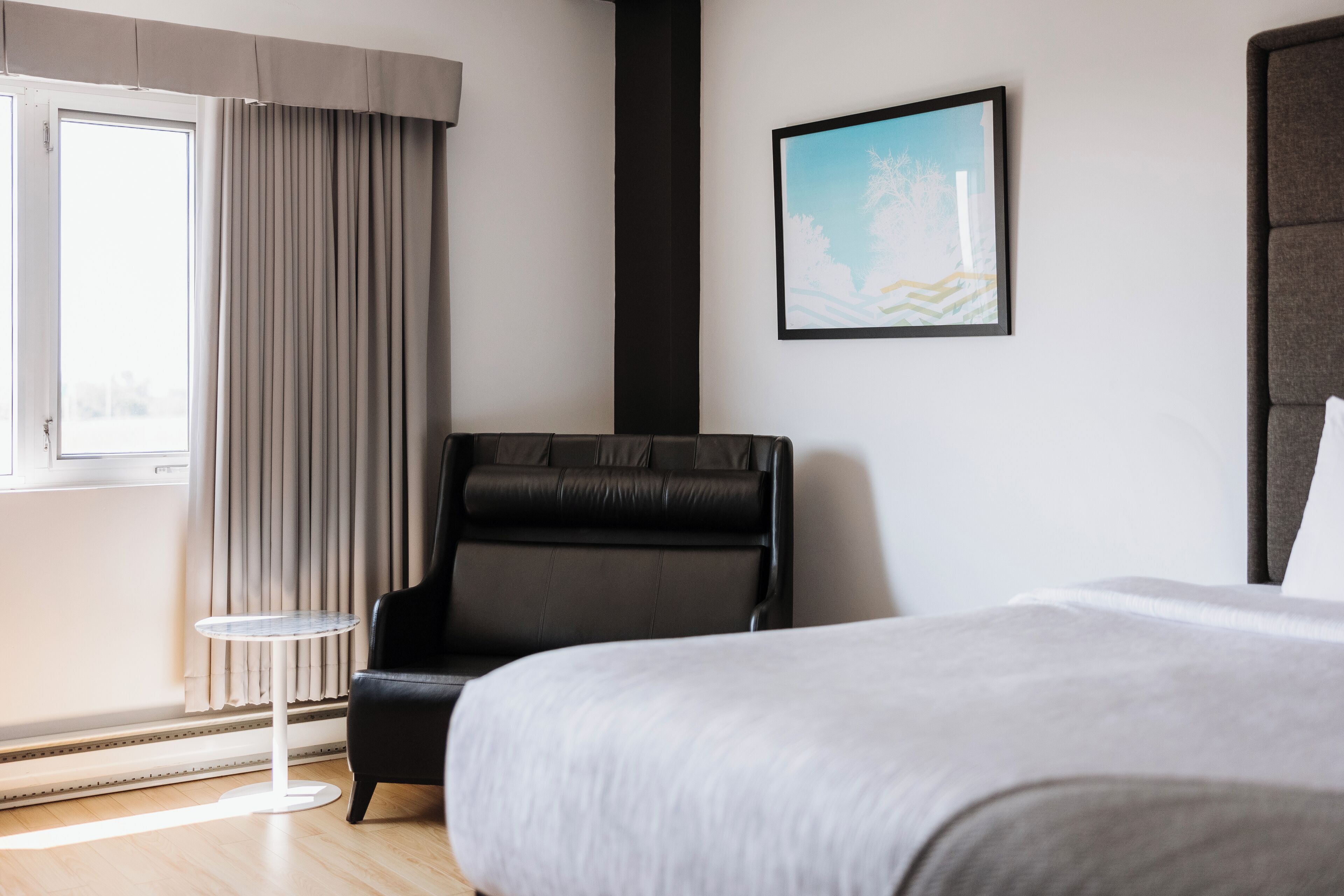 A hotel room with a bed, a black leather chair, a small round table, a window with curtains, and a framed picture on the wall.