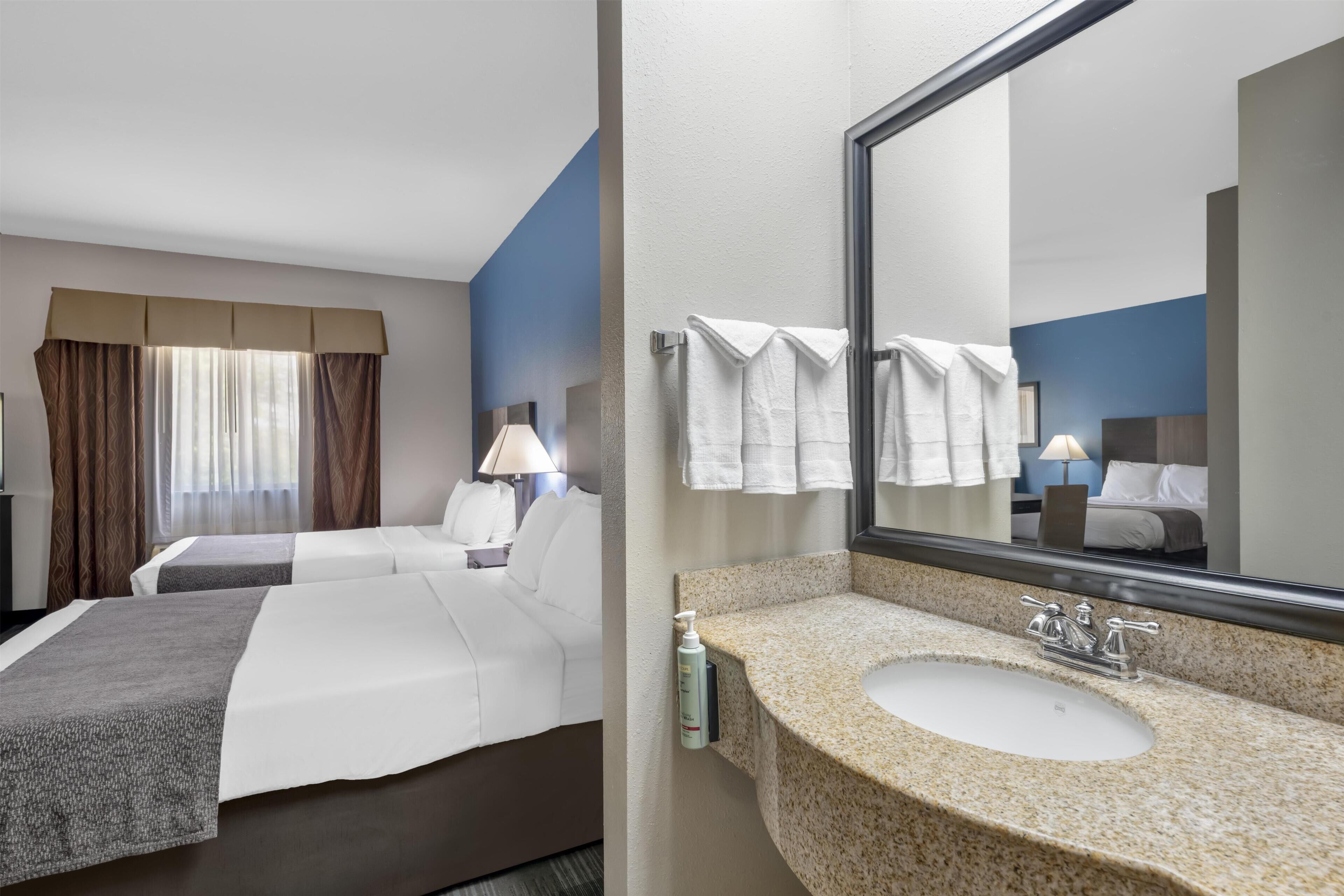 Ein Hotelzimmer mit zwei Betten, einem großen Spiegel, einem Waschbecken mit Granitplatte und einem Fenster mit Vorhängen.