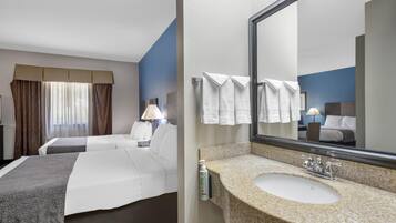 Ein Hotelzimmer mit zwei Betten, einem großen Spiegel, einem Waschbecken mit Granitplatte und einem Fenster mit Vorhängen.