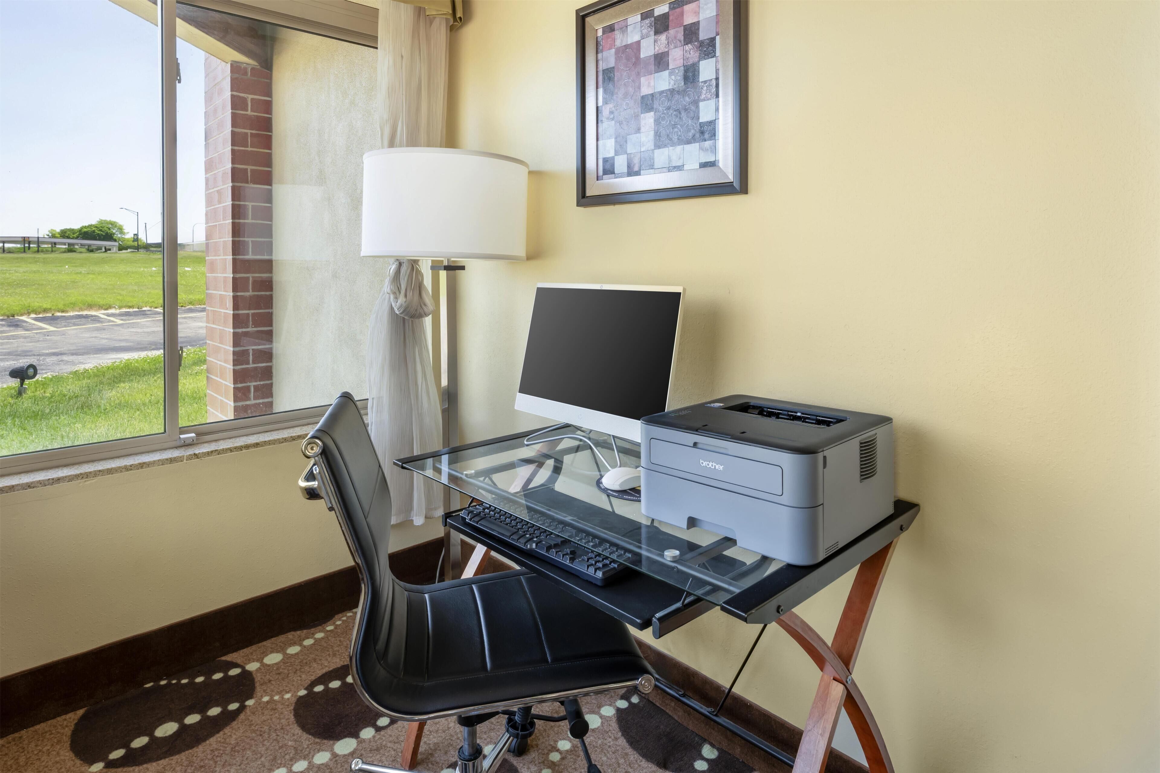 Habitación de hotel con escritorio de vidrio, computadora, impresora, silla, lámpara y ventana con vista a un estacionamiento.