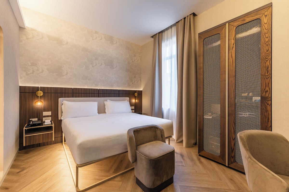 A hotel room with a large bed, a wooden nightstand, a chair, and a wardrobe with a mesh door.