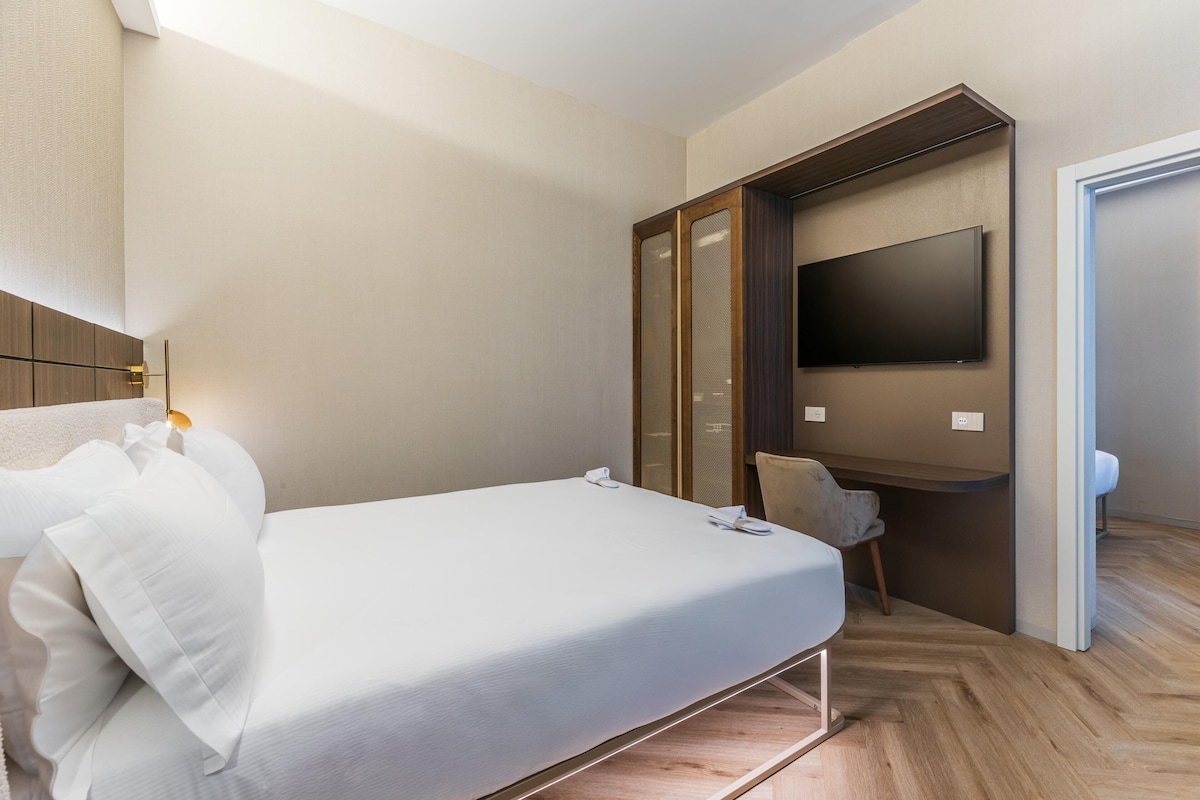 A hotel room with a bed, a TV mounted on the wall, a wardrobe, and a desk.