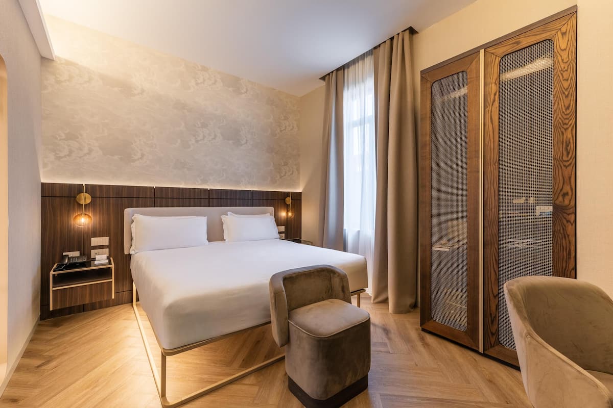 Una camera d'albergo con un letto grande, un comodino in legno, una sedia e un armadio con sportello a rete.