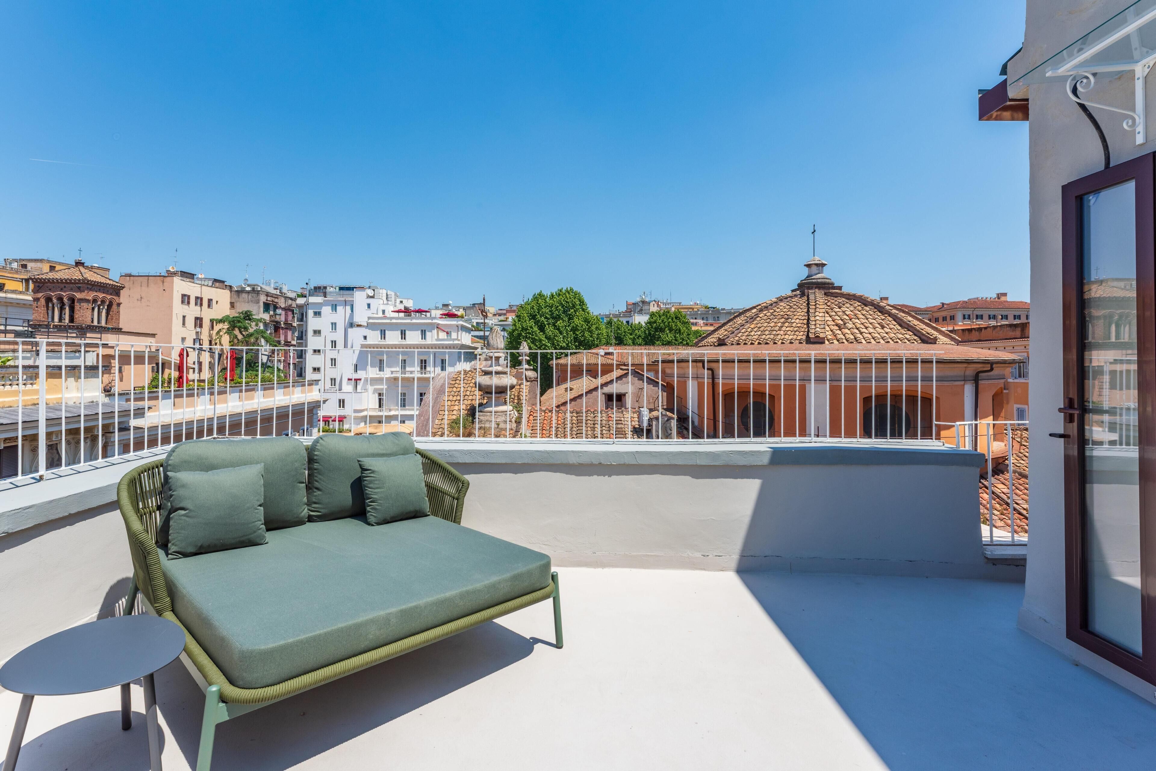 A balcony with a green sofa and a small table, overlooking a cityscape with buildings and a clear sky.