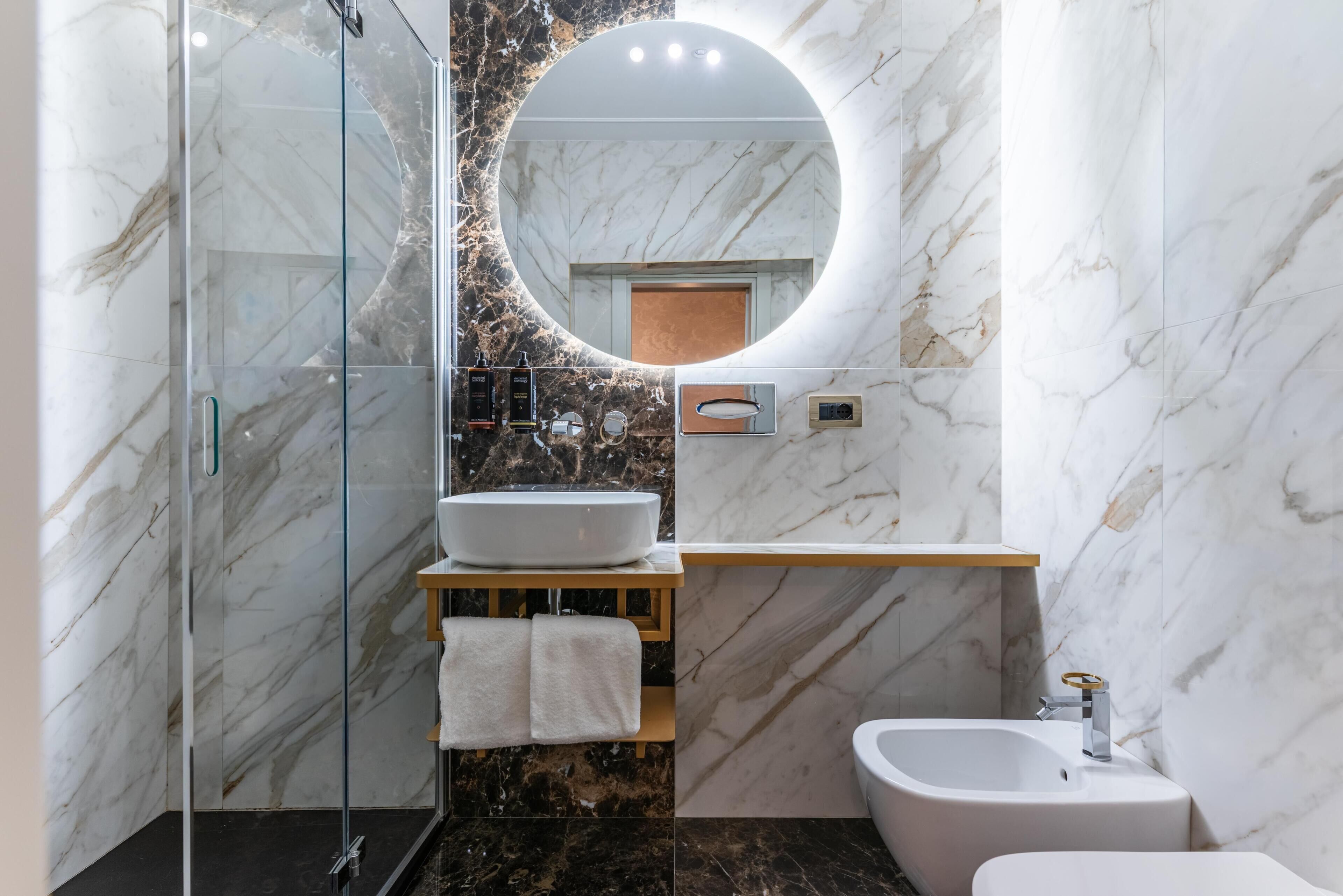 A modern bathroom with a glass shower enclosure, a marble-tiled wall, a round mirror, and a white sink.