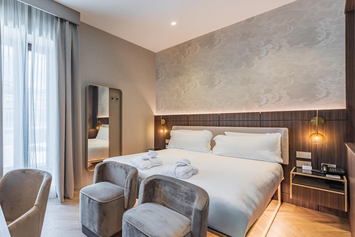 A modern hotel room with a large bed, two armchairs, a bedside table with a lamp, and a large mirror.
