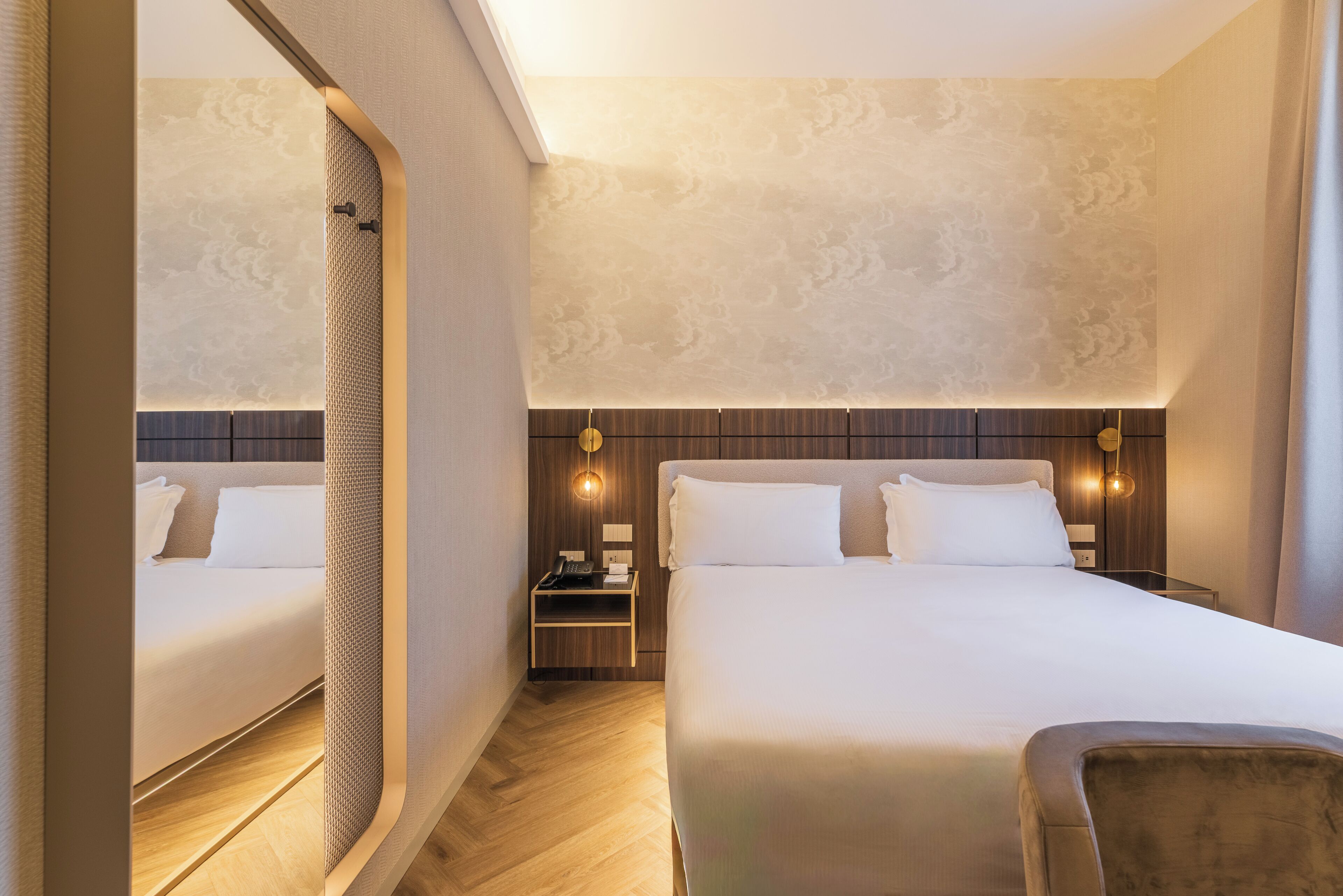 A hotel room with two beds, a wooden headboard, a nightstand with a lamp, and a mirror.
