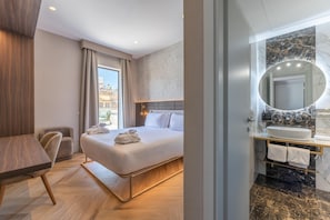 Quarto de hotel moderno com cama, escrivaninha, cadeira e banheiro com pia e espelho de mármore.