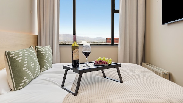 Quarto de hotel com uma cama, uma mesinha com uma garrafa de vinho, um copo de vinho e uma tigela de uvas.