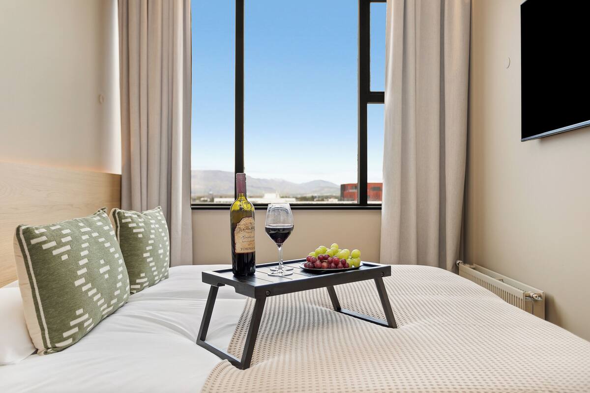 A hotel room with a bed, a small table with a bottle of wine, a glass of wine, and a bowl of grapes.