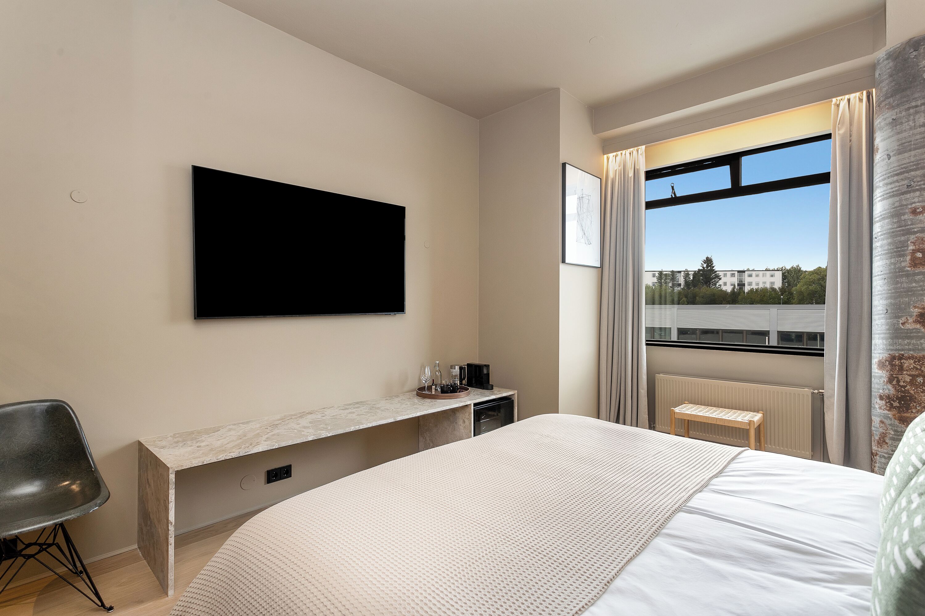 Una habitación de hotel moderna con una cama grande, un televisor de pantalla plana montado en la pared, un escritorio pequeño con una silla y una ventana con vistas a edificios y árboles.
