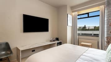 Una habitación de hotel moderna con una cama grande, un televisor de pantalla plana montado en la pared, un escritorio pequeño con una silla y una ventana con vistas a edificios y árboles.