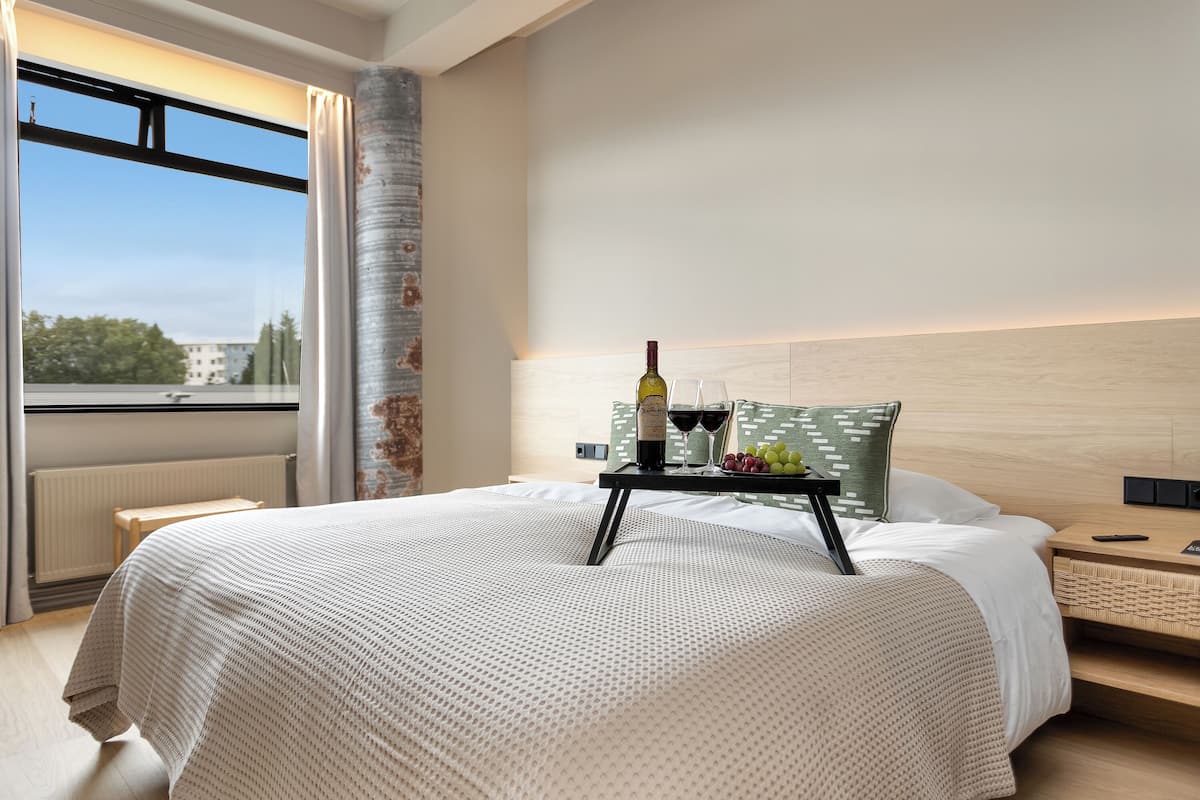 A modern hotel room with a large bed, a bedside table with a bottle of wine and glasses, and a view of the city through the window.