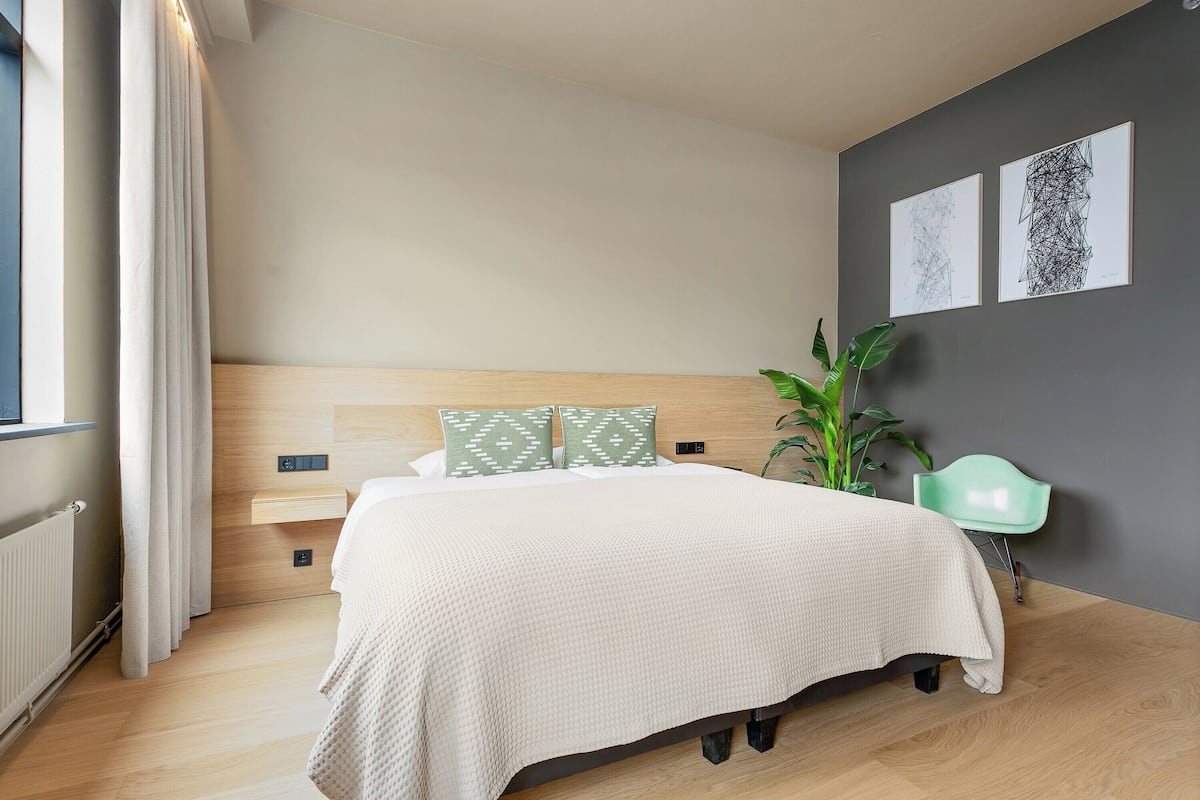 A modern bedroom with a large bed, a bedside table, a green chair, and two wall art pieces.