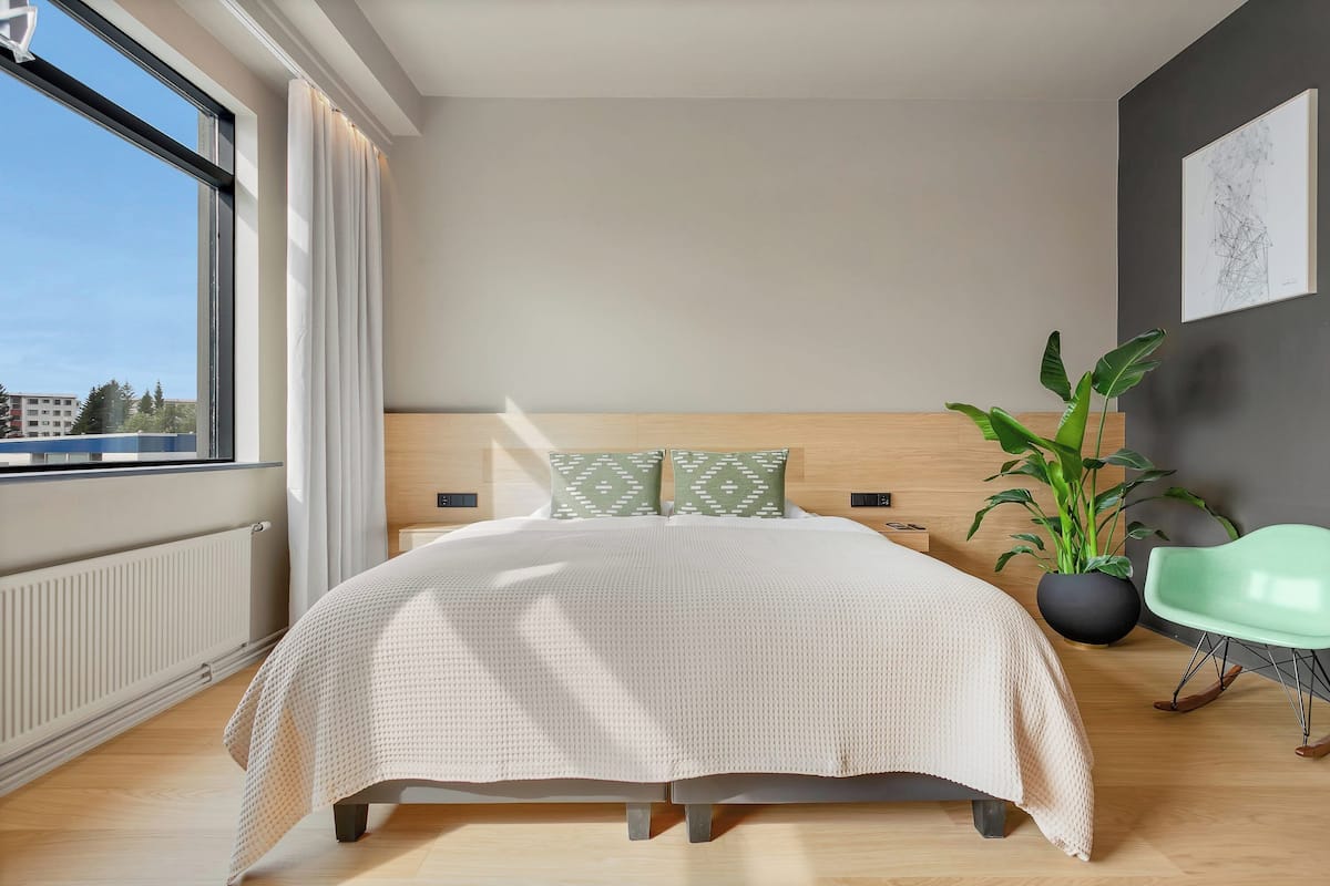 A modern bedroom with a large window, a bed with a beige blanket, a green plant, and a green chair.