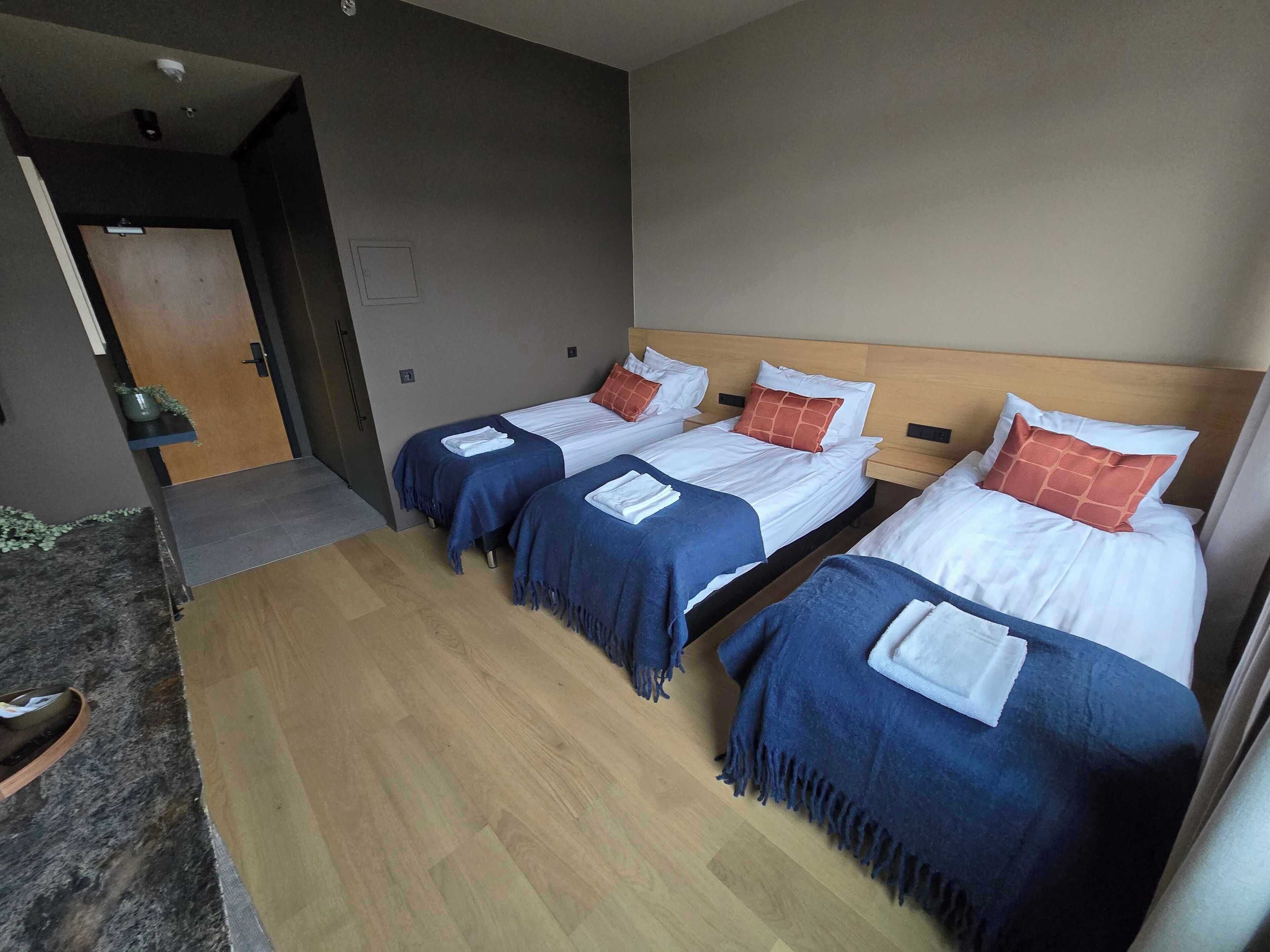 A hotel room with three beds, each with a blue bedspread and white linens. There is a wooden door and a small plant on the left side.