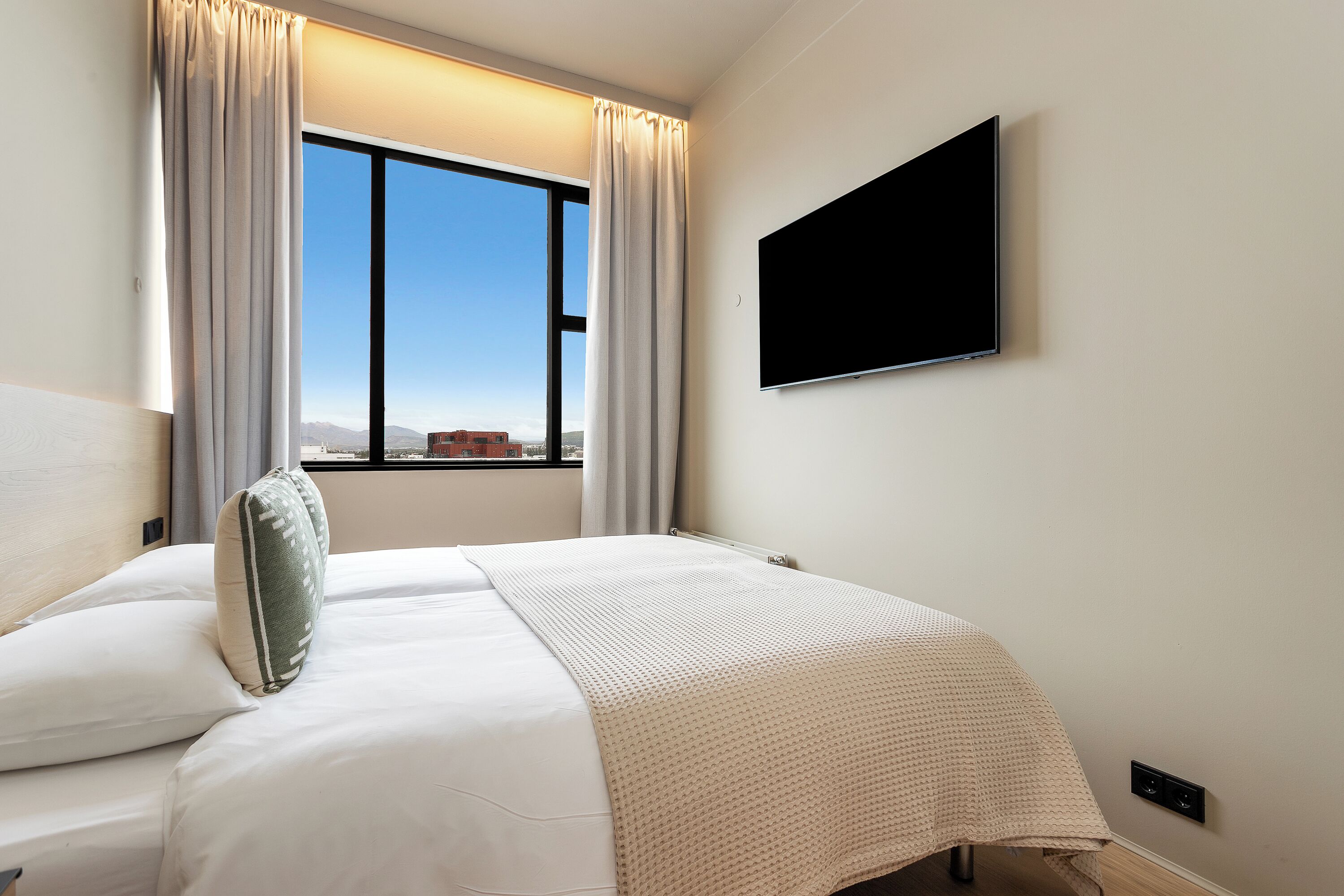 A hotel room with a large window, a bed with white bedding, a flat-screen TV mounted on the wall, and a view of the cityscape.