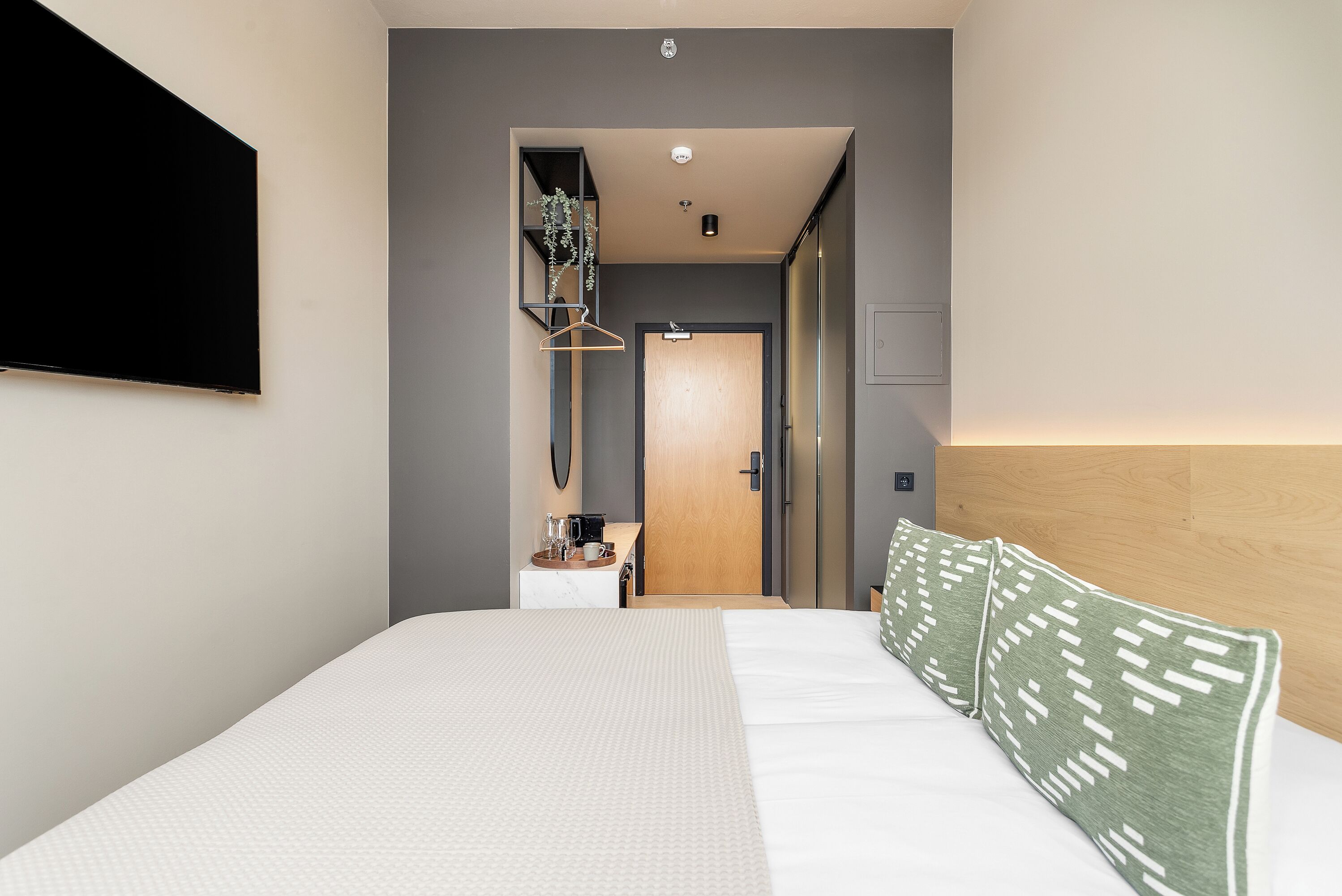 Una habitación de hotel moderna con una cama grande, un televisor de pantalla plana montado en la pared, un escritorio pequeño con una cafetera y una puerta de madera que conduce a otra habitación.