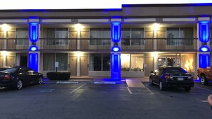 Un hotel de varios pisos con estacionamiento, que presenta una entrada central con columnas iluminadas y dos coches estacionados al frente.