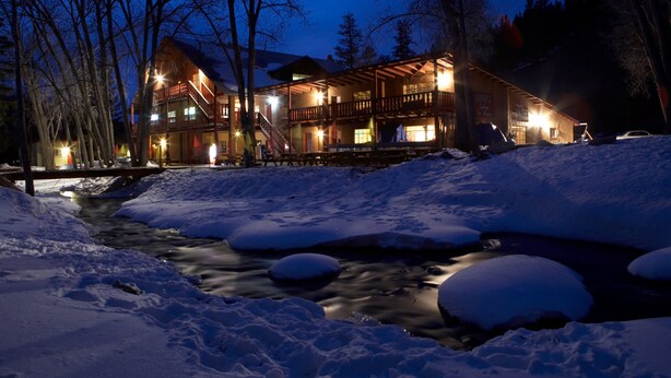 A lodge with a wooden exterior and multiple balconies, surrounded by snow and a flowing stream.