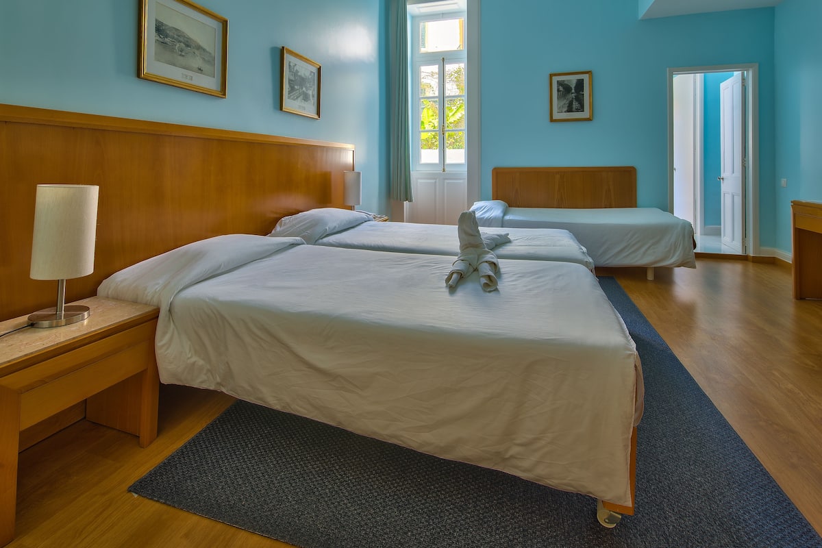 Une chambre d’hôtel avec deux lits, une tête de lit en bois, une table de chevet avec une lampe et une grande fenêtre avec des rideaux.