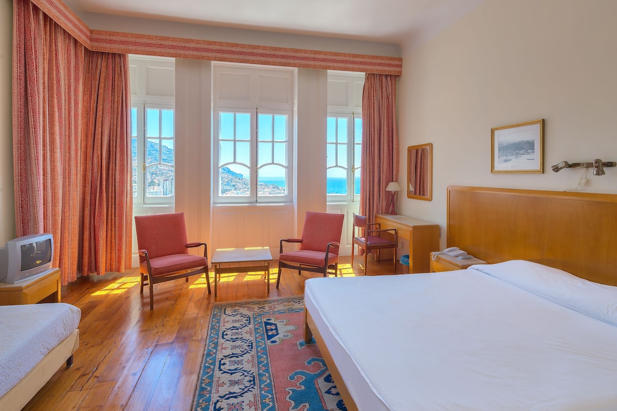 Une chambre d’hôtel comprenant un lit, deux fauteuils, un bureau, une télévision et une vue sur la mer grâce à de grandes fenêtres.