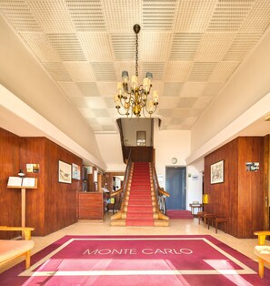 A hotel lobby with a red carpet reading "Monte Carlo," a grand staircase, wooden paneling, a chandelier, and a reception desk. - Hotel Monte Carlo (Funchal)