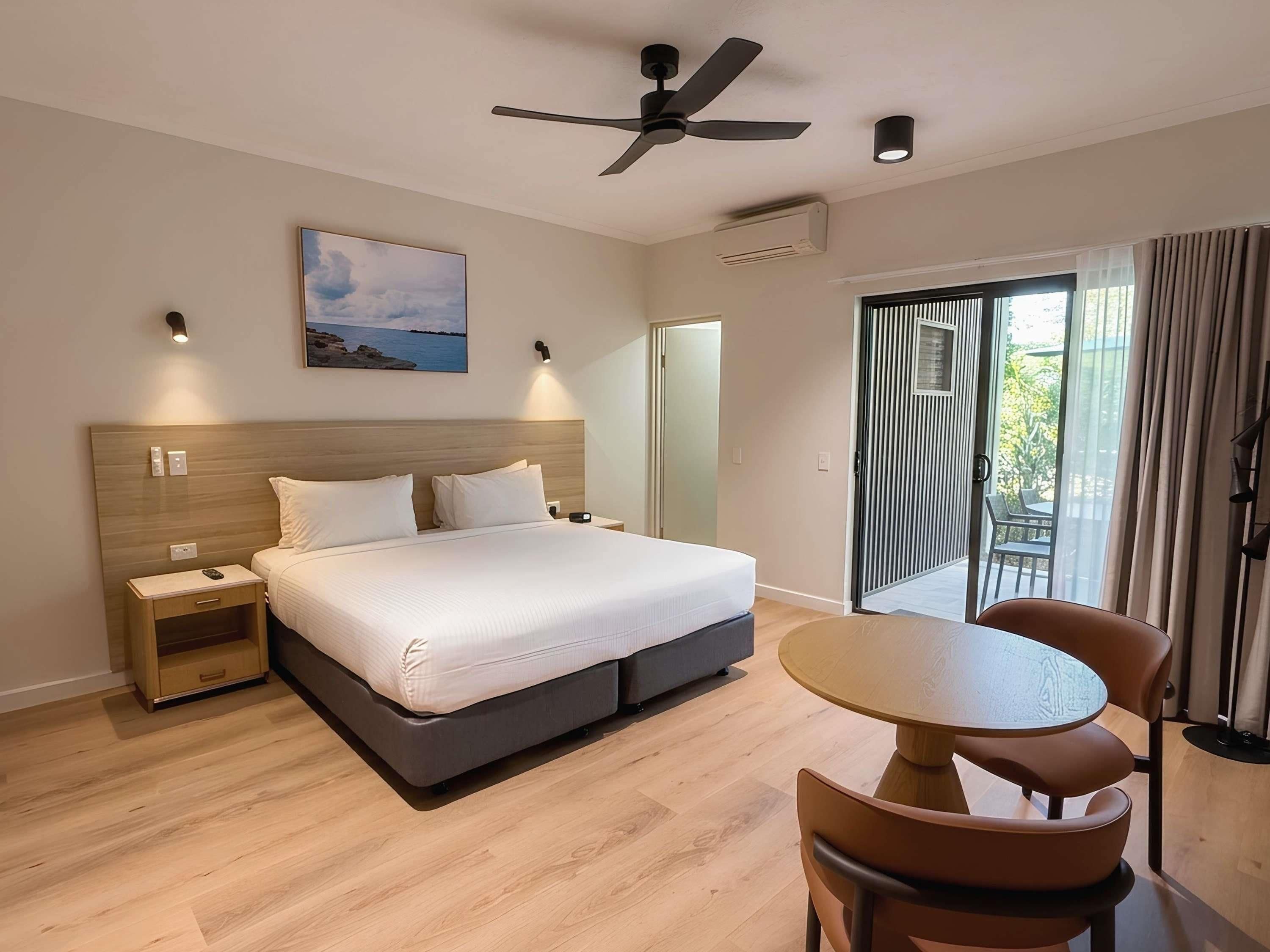 A hotel room with a bed, bedside tables, a ceiling fan, a painting, an air conditioning unit, a sliding glass door to a balcony, and a round table with chairs.