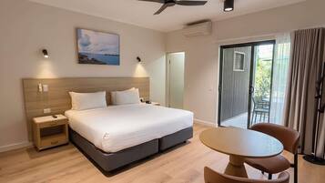 A hotel room with a bed, bedside tables, a ceiling fan, a painting, an air conditioning unit, a sliding glass door to a balcony, and a round table with chairs.