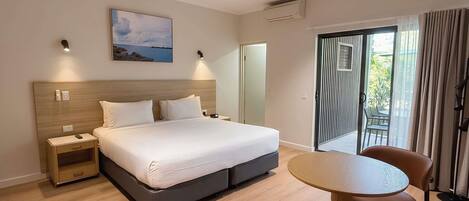 Habitación de hotel con cama, mesitas de noche, ventilador de techo, un cuadro, unidad de aire acondicionado, puerta corrediza de vidrio a un balcón y una mesa redonda con sillas.