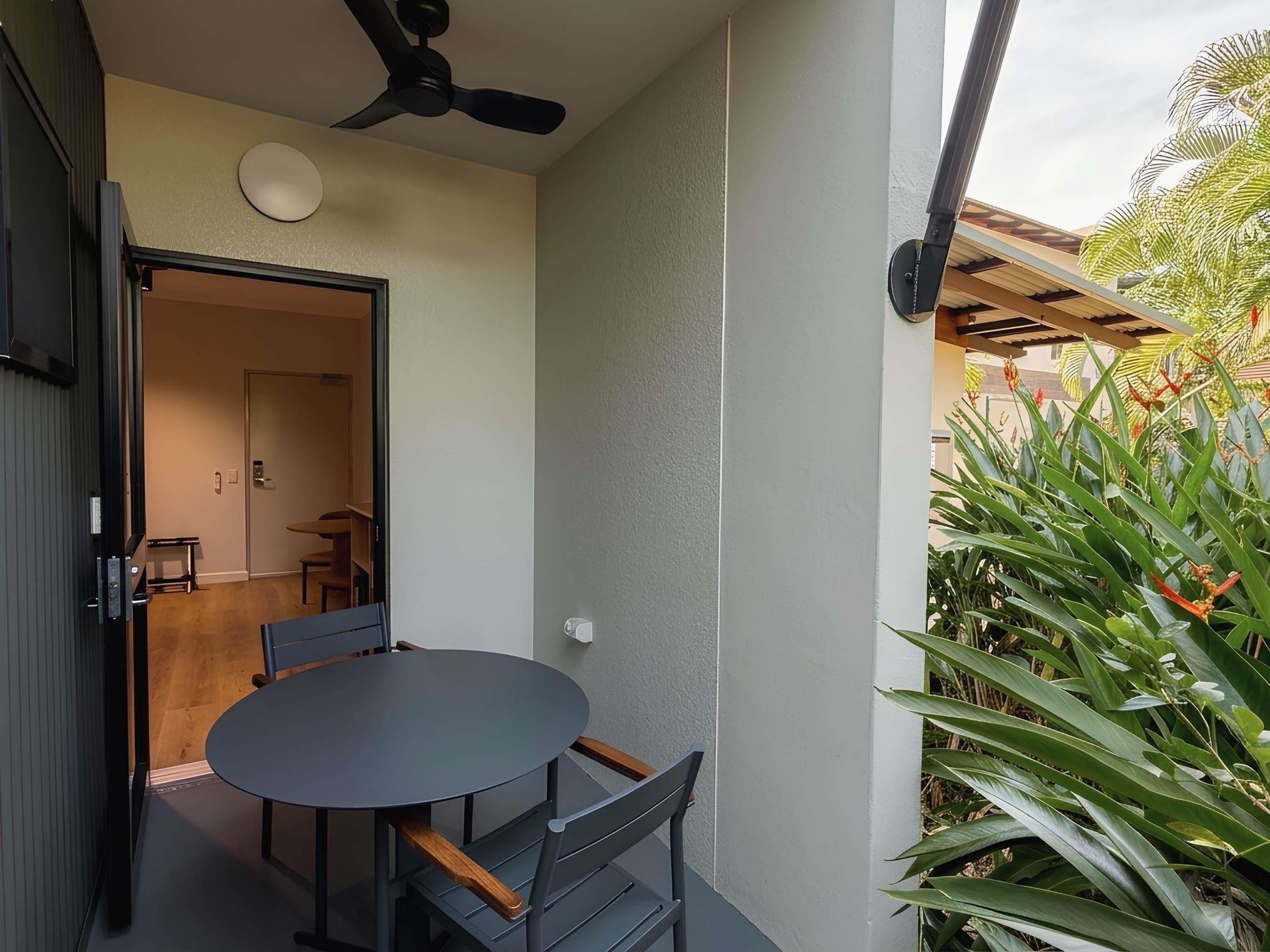 Un pequeño patio con una mesa y sillas, un ventilador de techo y una puerta que da a un espacio interior.